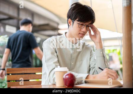 Un jeune homme asiatique concentré et sérieux contemple et note ses idées dans un livre tout en étant assis à une table en plein air dans un café. people et li Banque D'Images