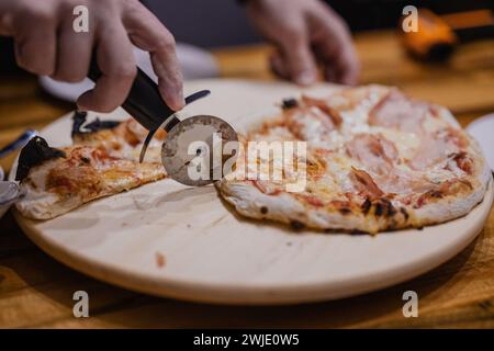 Personne coupant une pizza faite maison avec une lame de rouleau. Pizza croustillante fraîchement sortie du four sur une assiette en bois. Mains d'une personne vue le couper Banque D'Images