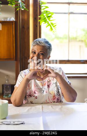 Une femme biraciale mature est joyeuse, faisant une forme de cœur avec ses mains à la maison sur un appel vidéo Banque D'Images