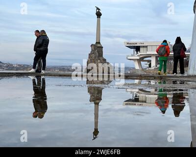 Sewastopol, Ukraine. 13 février 2024. Les gens se promènent le long du boulevard côtier de la ville portuaire devant le monument aux navires coulés, qui se reflète dans une flaque d'eau. C’est un rappel que l’Empire russe a délibérément coulé des navires là-bas en 1854 afin de créer une barrière artificielle contre les envahisseurs de la mer. Cette année, la Russie célèbre le 10e anniversaire de l’annexion de la Crimée, qui fait partie de l’Ukraine en vertu du droit international. Crédit : Ulf Mauder/dpa/Alamy Live News Banque D'Images