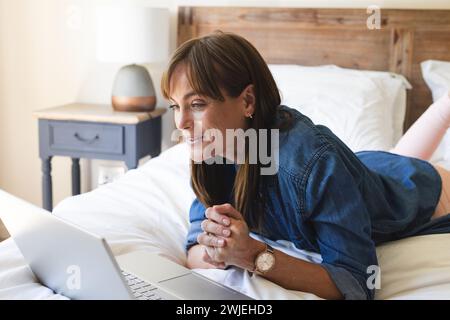 Une femme caucasienne d'âge moyen sourit tout en utilisant un ordinateur portable dans un cadre de chambre confortable Banque D'Images