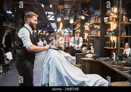 Client régulier visitant le salon de coiffure préféré. Coiffeur professionnel et client souriant. Barbier masculin portant une cape blanche. Banque D'Images