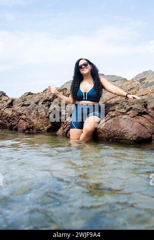 Salvador, bahia, Brésil - 06 janvier 2024 : belle femme portant des lunettes de soleil appuyées contre des rochers sur une plage. Personne heureuse voyageant. Banque D'Images