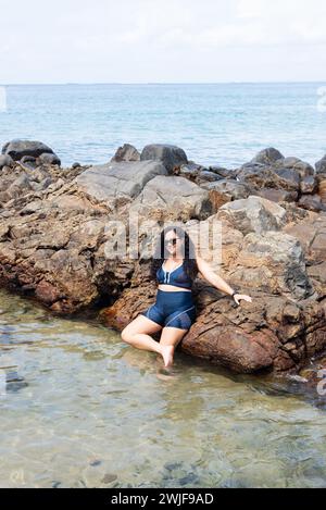 Salvador, bahia, Brésil - 06 janvier 2024 : belle femme portant des lunettes de soleil appuyées contre des rochers sur une plage. Personne heureuse voyageant. Banque D'Images