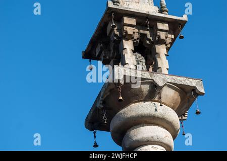 Une photo détaillée capturant l'architecture en pierre ornée d'un pilier sculpté à Shravanabelagola, avec de petites cloches suspendues posées sur un ciel bleu. Banque D'Images