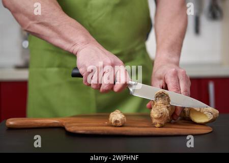 Homme cuisinier peler et nettoyer une grande racine de gingembre sur une planche de bois Banque D'Images