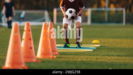 Garçon de football tenant la balle dans les mains et sautant par-dessus le sentier des haies. Athlétisme Agility Hurdles Track. Garçon améliorant les compétences de vitesse et d'agilité Banque D'Images