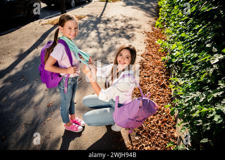 Pleine longueur au-dessus de la vue portrait d'une jeune fille joyeuse nouant mouchoir la petite sœur porter sac à dos école la lumière du soleil à l'extérieur Banque D'Images