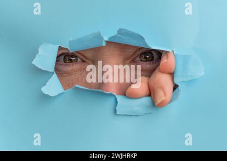 Femme jetant un coup d'œil par un trou dans un panneau d'affiche bleu déchiré, sa main est visible Banque D'Images