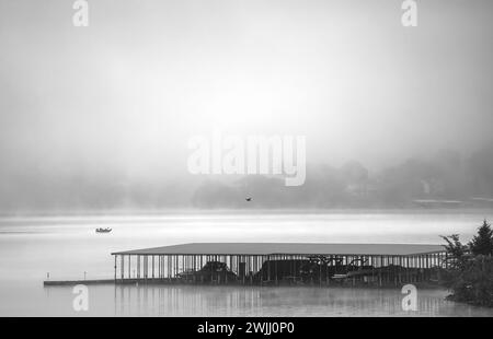 Bateau de pêche voyageant à travers un lac calme sur un matin brumeux devant un quai de bateau avec un oiseau noir volant à travers la scène en noir et blanc. Banque D'Images