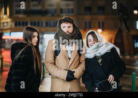 Trois amis apprécient la compagnie de l'autre lors d'une nuit hivernale, enveloppés dans des manteaux chauds et des foulards, mettant en valeur l'amitié et la vie urbaine. Banque D'Images
