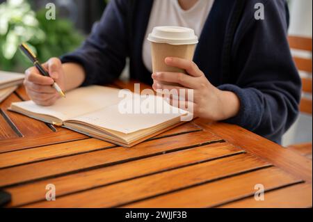 Photo recadrée d'une femme en vêtements décontractés sirotant un café et écrivant ses idées dans un livre alors qu'elle était assise dans un café. tenir un journal, noter, Banque D'Images