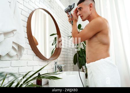 Jeune homme attrayant séchant ses cheveux après la douche dans la salle de bain Banque D'Images