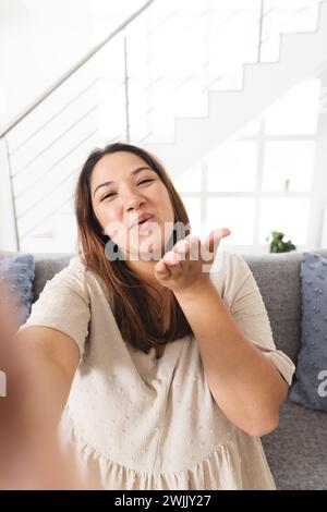 Jeune femme biraciale souffle un baiser à l'intérieur sur un appel vidéo, avec un espace de copie Banque D'Images