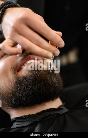 Un coiffeur styliste applique des gouttes d'huile sur la barbe des clients pour hydrater et adoucir. Soin de la barbe. Banque D'Images