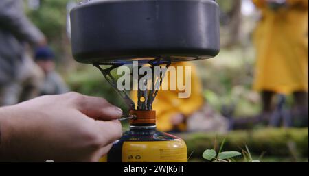 Bouilloire touristique se tient sur la cuisinière à gaz portable de camping. Un voyageur masculin allume un brûleur à gaz. Groupe de copains de randonnée s'est arrêté pour se reposer après trek dans les montagnes. Concept de découverte de la nature et de loisirs actifs. Banque D'Images