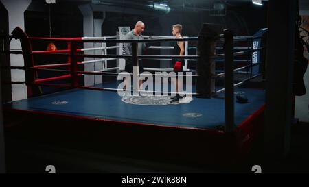 Garçon dans des gants de boxe se tient sur l'anneau et parle avec l'entraîneur. Homme adulte consulte jeune boxeur et explique les techniques de combat. Adolescent se prépare à un match de combat dans la salle de gym sombre. Activité physique et entraînement. Banque D'Images