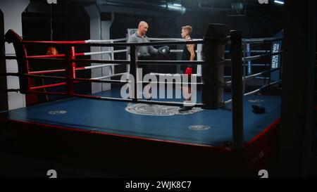 Garçon dans des gants de boxe se tient sur l'anneau et parle avec l'entraîneur. Homme adulte consulte jeune boxeur et explique les techniques de combat. Adolescent se prépare à un match de combat dans la salle de gym sombre. Activité physique et entraînement. Banque D'Images