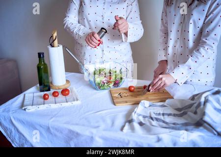 adolescente et sa mère en pyjama cuisinent et mangent une salade verte fraîche et de tomates ensemble Banque D'Images