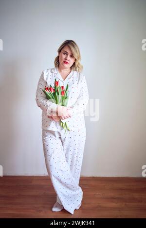 un portrait d'une belle femme de taille moyenne en pyjama avec un bouquet de tulipes rouges dans un style minimaliste Banque D'Images