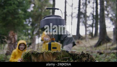 Bouilloire touristique se tient sur la cuisinière à gaz portable de camping. Un voyageur masculin allume un brûleur à gaz. Groupe de copains de randonnée s'est arrêté pour se reposer après trek dans les montagnes. Concept de découverte de la nature et de loisirs actifs. Banque D'Images