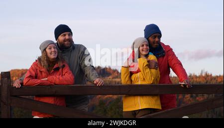 Les couples multiethniques se tiennent près d'une clôture en bois, regardent le beau coucher de soleil en profitant de la nature pendant le voyage. Groupe de touristes ou de copains de randonnée se reposent après la randonnée dans les montagnes. Passionnés d'activités de plein air. Ralenti. Banque D'Images