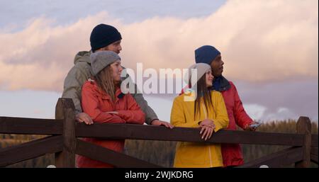 Les couples multiethniques se tiennent près d'une clôture en bois, regardent le beau coucher de soleil en profitant de la nature pendant le voyage. Groupe de touristes ou de copains de randonnée se reposent après la randonnée dans les montagnes. Passionnés d'activités de plein air. Ralenti. Banque D'Images