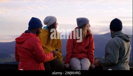 Les copains de randonnée se tiennent près d'une clôture en bois et regardent le coucher du soleil. Groupe de divers touristes ou voyageurs apprécient le paysage des montagnes et la belle nature pendant le voyage. Passionnés d'activités de plein air. Ralenti. Banque D'Images