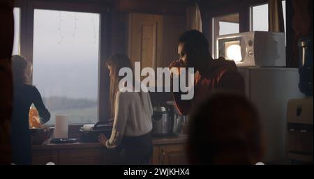 Groupe multiethnique d'amis cuisinent le petit déjeuner ensemble dans la maison de vacances. Homme afro-américain se tient dans la cuisine, boit du jus. La famille des grands voyageurs s'est arrêtée pour se reposer dans le chalet pendant les vacances. Tourisme. Banque D'Images