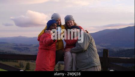 Les copains de randonnée se tiennent près d'une clôture en bois et regardent le coucher du soleil. Groupe de divers touristes ou voyageurs apprécient le paysage des montagnes et la belle nature pendant le voyage. Passionnés d'activités de plein air. Ralenti. Banque D'Images