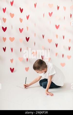 Jeune garçon dessinant un coeur sur papier blanc par fond de guirlande de coeur Banque D'Images