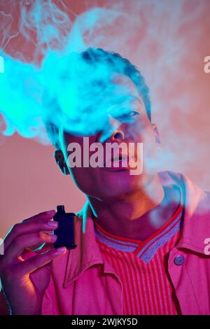 portrait de mec afro-américain exhalant de la fumée tout en tenant e cigarette sur fond vibrant Banque D'Images