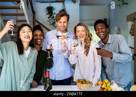 Groupe d'amis regardant la caméra griller avec des verres à vin debout dans la cuisine Banque D'Images