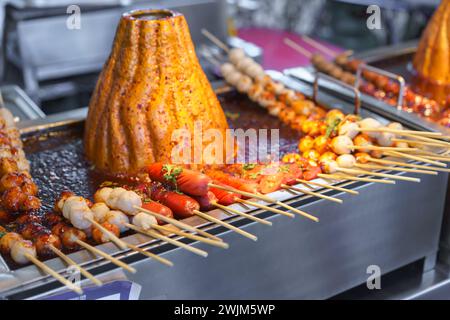 Boule de porc en brochettes comprenant des saucisses cuites sur un grill chaud, arrosées d'une sauce épicée, dans un marché alimentaire de rue. Banque D'Images