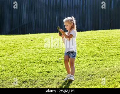 Petite fille debout sur la pelouse dans le parc de la ville d'été et tenant smartphone. Concept d'enfance, de loisirs et de personnes - repos heureux de l'enfant et avoir un g Banque D'Images