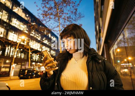 Jeune belle femme noire tapant un message en ligne sur son téléphone tout en marchant dans la rue d'une ville. Banque D'Images