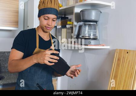 Jeune homme latin, concentré, heureux à la cuisine à la maison, préparant le moule pour verser le mélange de gâteau, en utilisant le moule rectangulaire en téflon. Banque D'Images