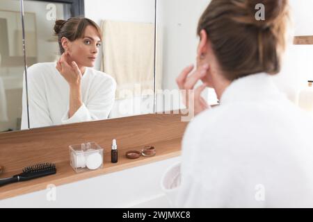 Une femme caucasienne d'âge moyen examine sa peau dans un miroir de salle de bain Banque D'Images