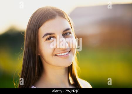 Portrait d'une femme brune blanche dans la vingtaine, à la lumière du soleil couchant sur fond flou de paysage rural, gros plan. Banque D'Images