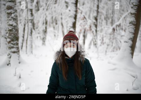 une femme debout au milieu d'une forêt enneigée, portant un masque blanc, une veste d'hiver sarcelle et un chapeau multicolore. Les arbres autour d'elle sont le dr. Banque D'Images