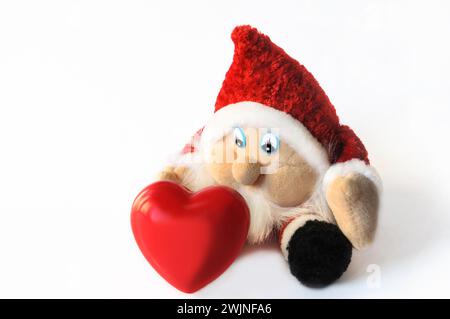 Un gnome jouet dans des vêtements de Noël sur un fond blanc veut prendre un cœur décoratif. Banque D'Images