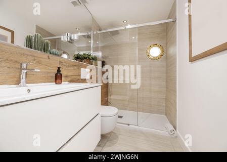 Image frontale d'une salle de bains avec des meubles blancs, mur carrelé avec des carreaux d'aspect bois, miroir à infini et cabine de douche avec hublot Banque D'Images