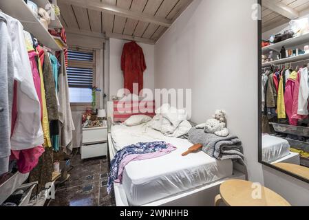 Très petite chambre avec un lit simple avec un grand désordre de vêtements sur le matelas, vêtements suspendus sur un cintre et plafonds avec langue et rainure woo Banque D'Images