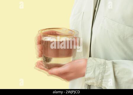 Femme tenant le verre d'eau dans les mains tremblantes sur fond jaune, gros plan. Le mois de la sensibilisation à la maladie de Parkinson Banque D'Images