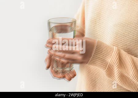 Femme tenant le verre d'eau dans les mains tremblantes sur fond clair, gros plan. Le mois de la sensibilisation à la maladie de Parkinson Banque D'Images