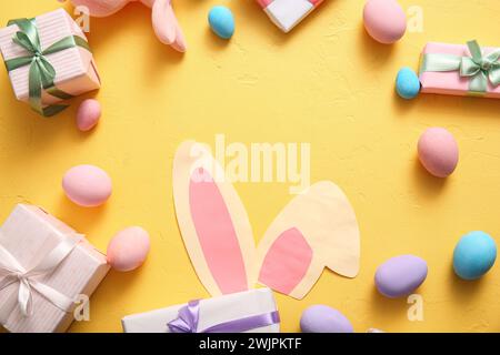 Cadre fabriqué à partir d'oreilles de lapin en papier, d'œufs de Pâques et de boîtes-cadeaux sur fond jaune Banque D'Images