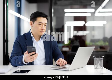 Un homme d'affaires asiatique professionnel travaille sur un ordinateur portable tout en vérifiant son smartphone dans un environnement de bureau moderne. Banque D'Images