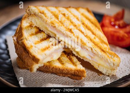 Sandwich toast coupé en tranches avec fromage Gouda fondu et jambon de dinde sur fond de bois avec tomate cerise sur une assiette noire. Servi avec du thé glacé. Banque D'Images