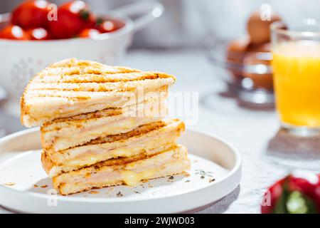 Sandwich toast coupé tranché avec fromage Gouda fondu jambon de dinde sur un fond lumineux avec des ingrédients jus d'orange tomate oeufs en fond flou Banque D'Images