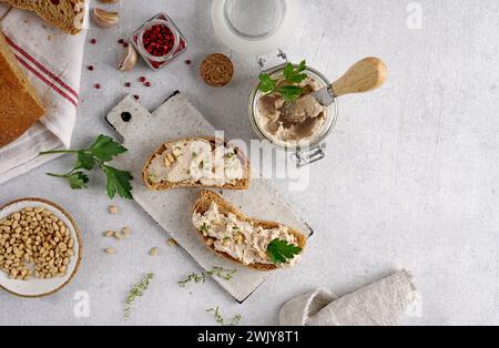 Pâté maison, tartinade ou mousse dans un pot en verre avec du pain tranché et des herbes avec des pignons de pin, fond de béton léger. Banque D'Images